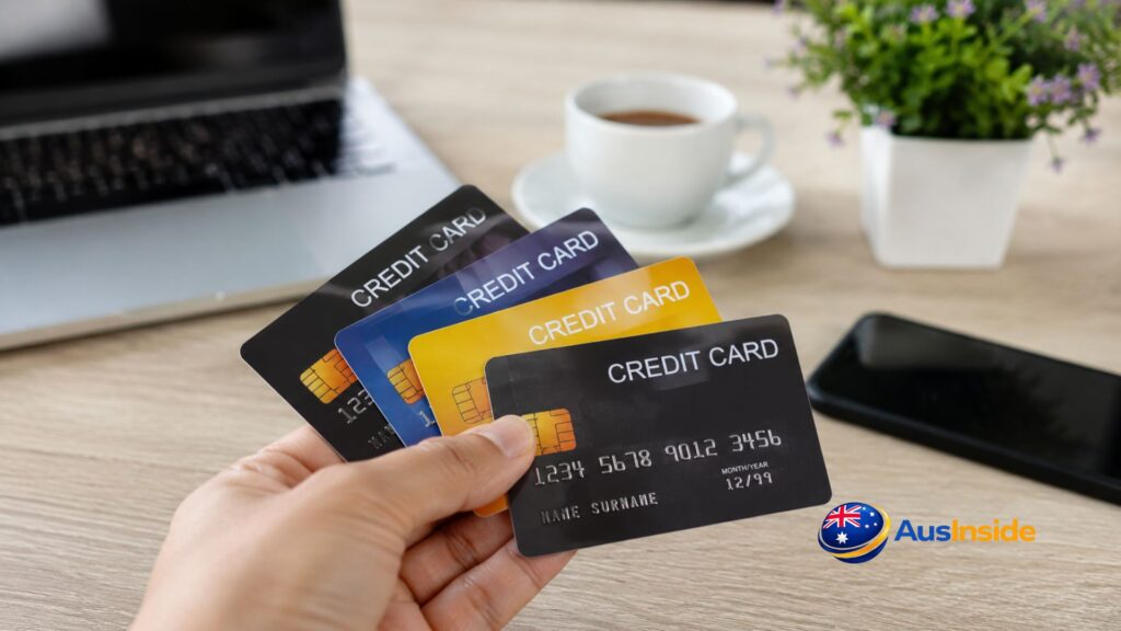Hand holding multiple credit cards above a desk with a laptop, coffee cup, smartphone, and plant in the background, representing credit card comparison and personal finance in Australia.