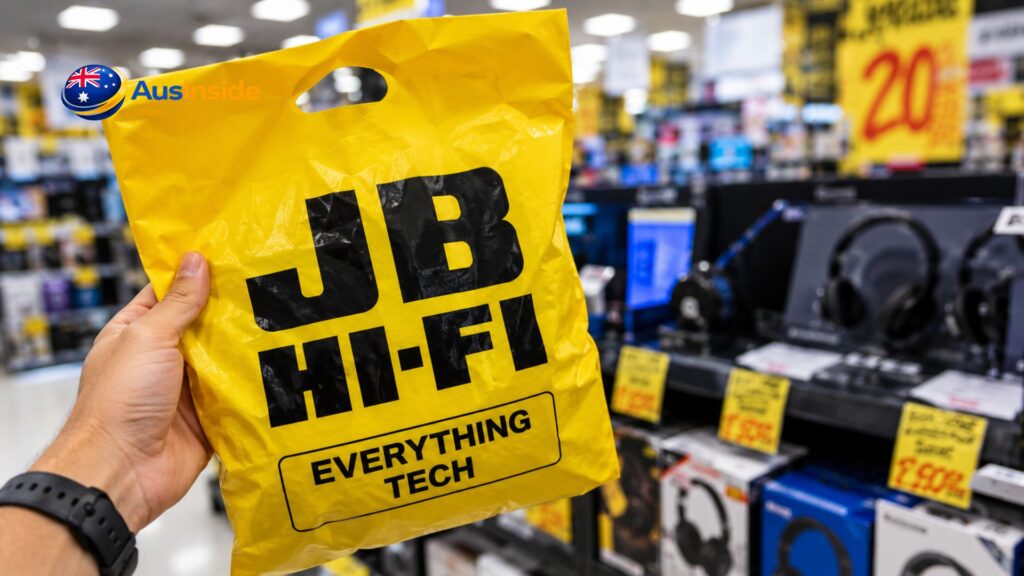 Customer holding a yellow JB Hi-Fi shopping bag inside an electronics store, with headphones, tech products, and promotional sale signage visible in the background.