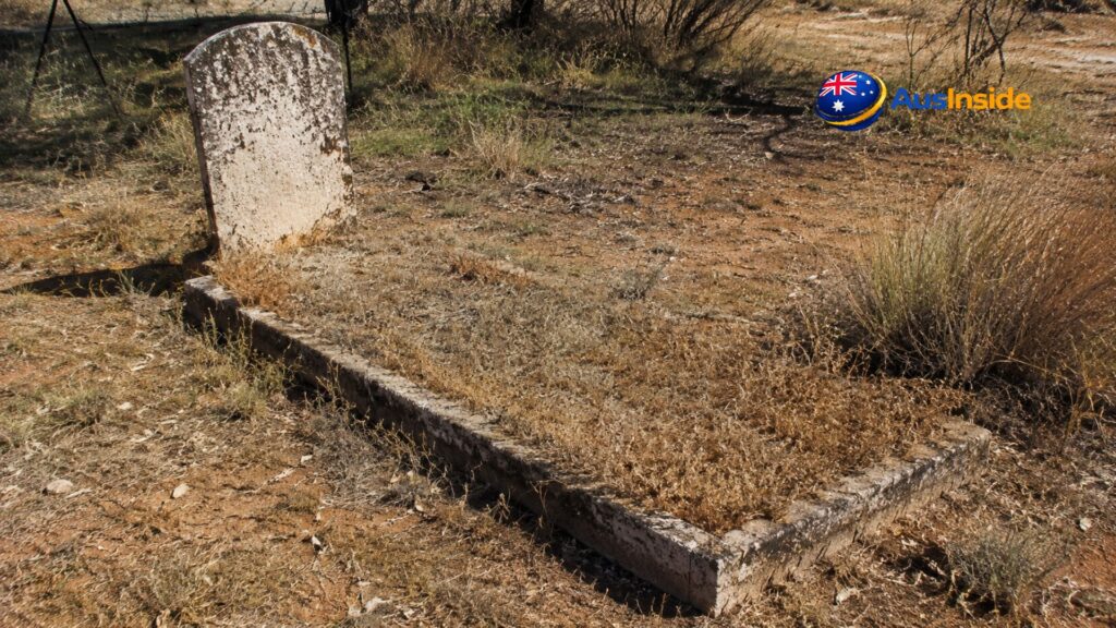 Weathered single grave with an aged white headstone surrounded by dry grass and red outback soil at Bedourie Cemetery, Queensland, Australia.