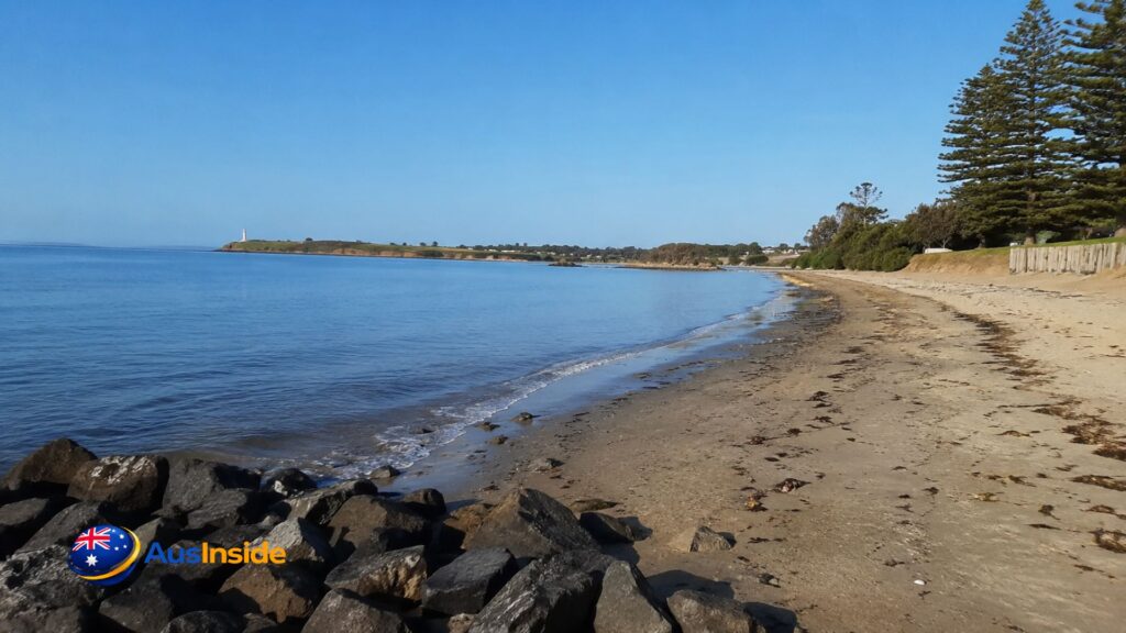 East Devonport Beach with calm waters, sandy shoreline, rocky foreground, and distant Mersey Bluff lighthouse under a clear blue sky in Tasmania.